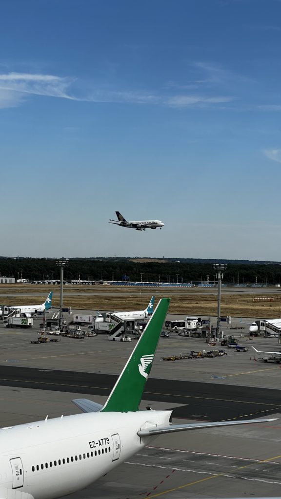 La queue d'un Boeing 777-22K(LR) de Turkmenistan Airlines immatriculé EZ-A779 à sa porte d'embarquement l’aéroport de Frankfurt (FRA) en Allemagne avec en arrière-plan un A380 de Singapore Airlines effectuant un atterrissage.